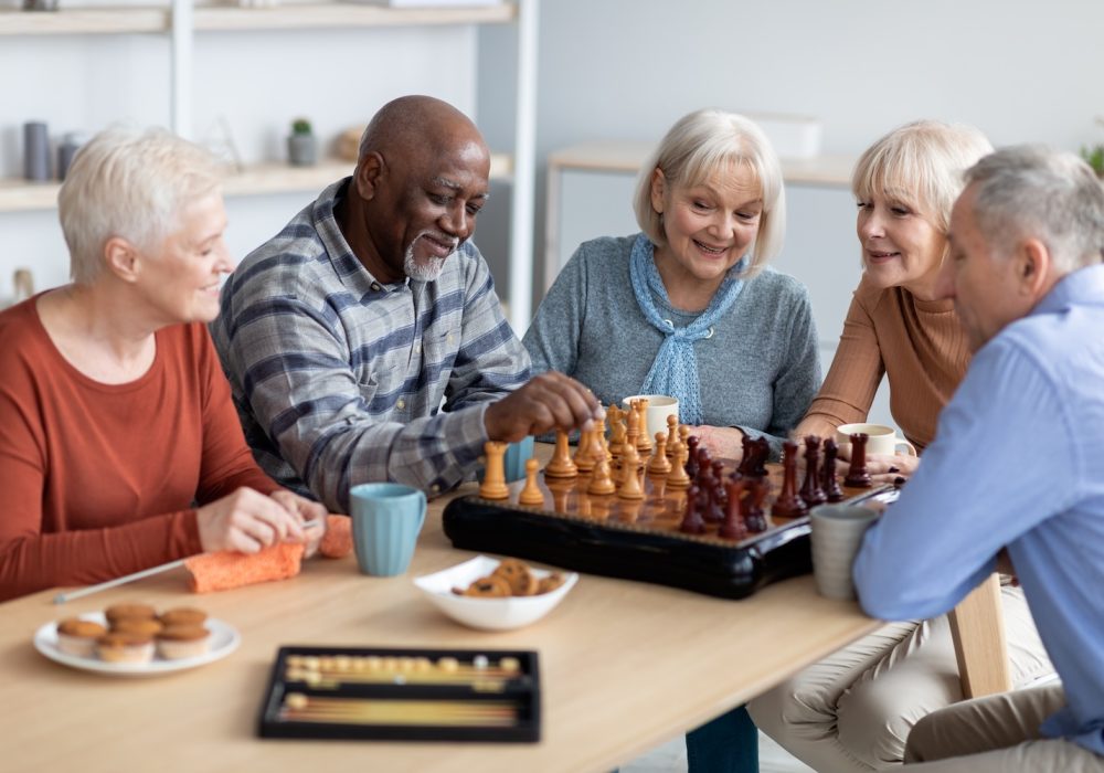 Multiracial senior people men and women in casual playing chess at nursing home, sitting around table, drinking tea with cookies, playing table games, knitting, spending time together